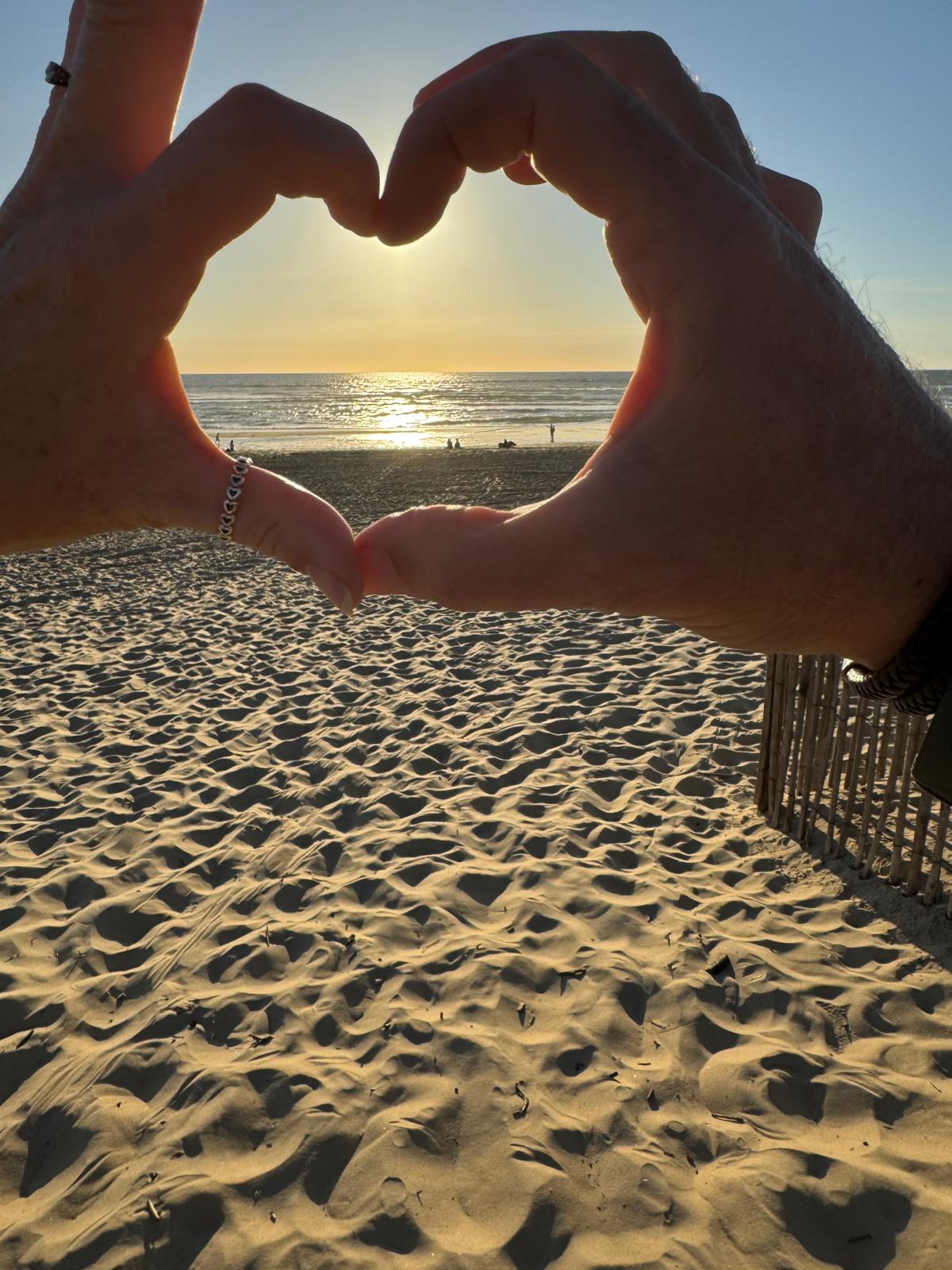 Two hands clasped into a heart framing the sun setting over the Atlantic surf at Messanges.