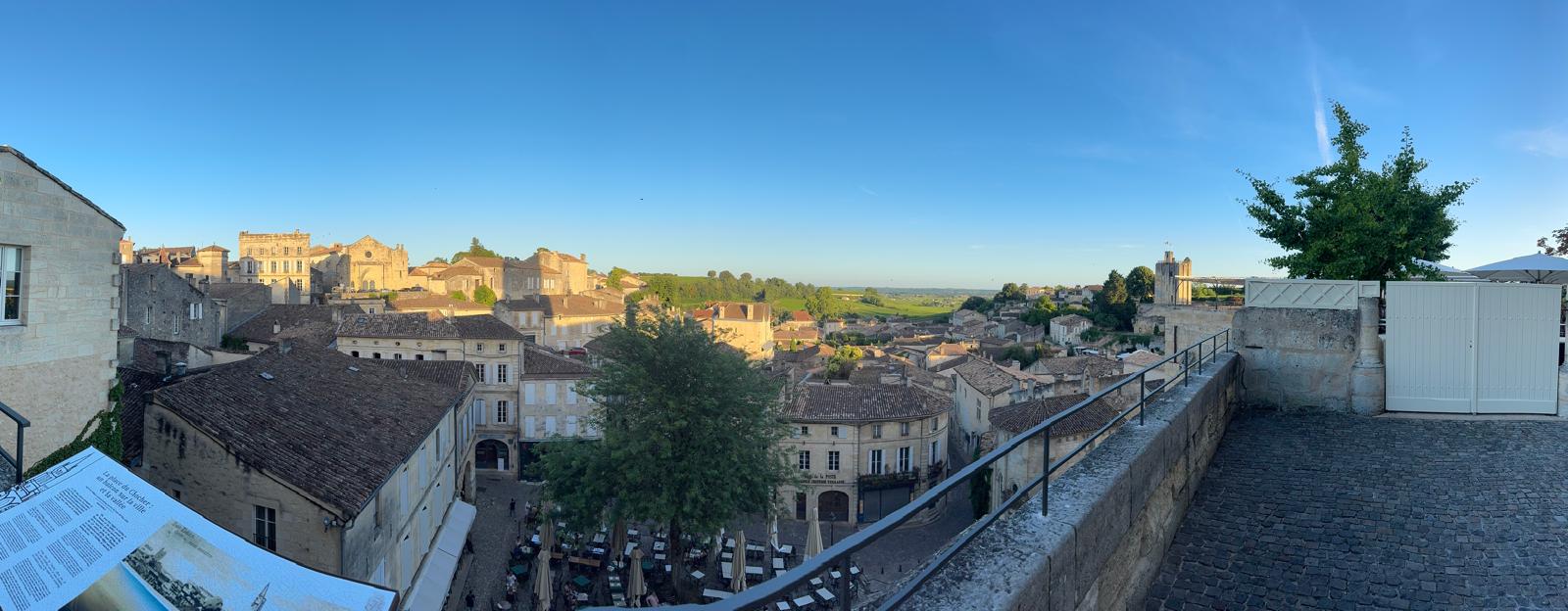 Golden-hour panorama over the rooftops of Saint-Émilion, the monolithic bell tower on the skyline and vineyards beyond.