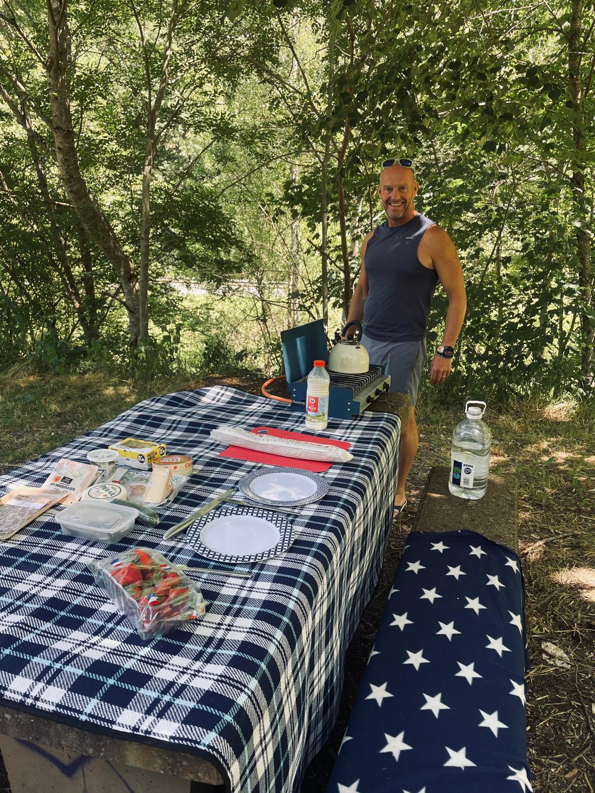 Dave at a picnic table laid with a checked cloth and camp kettle at a roadside aire in the High Velay.