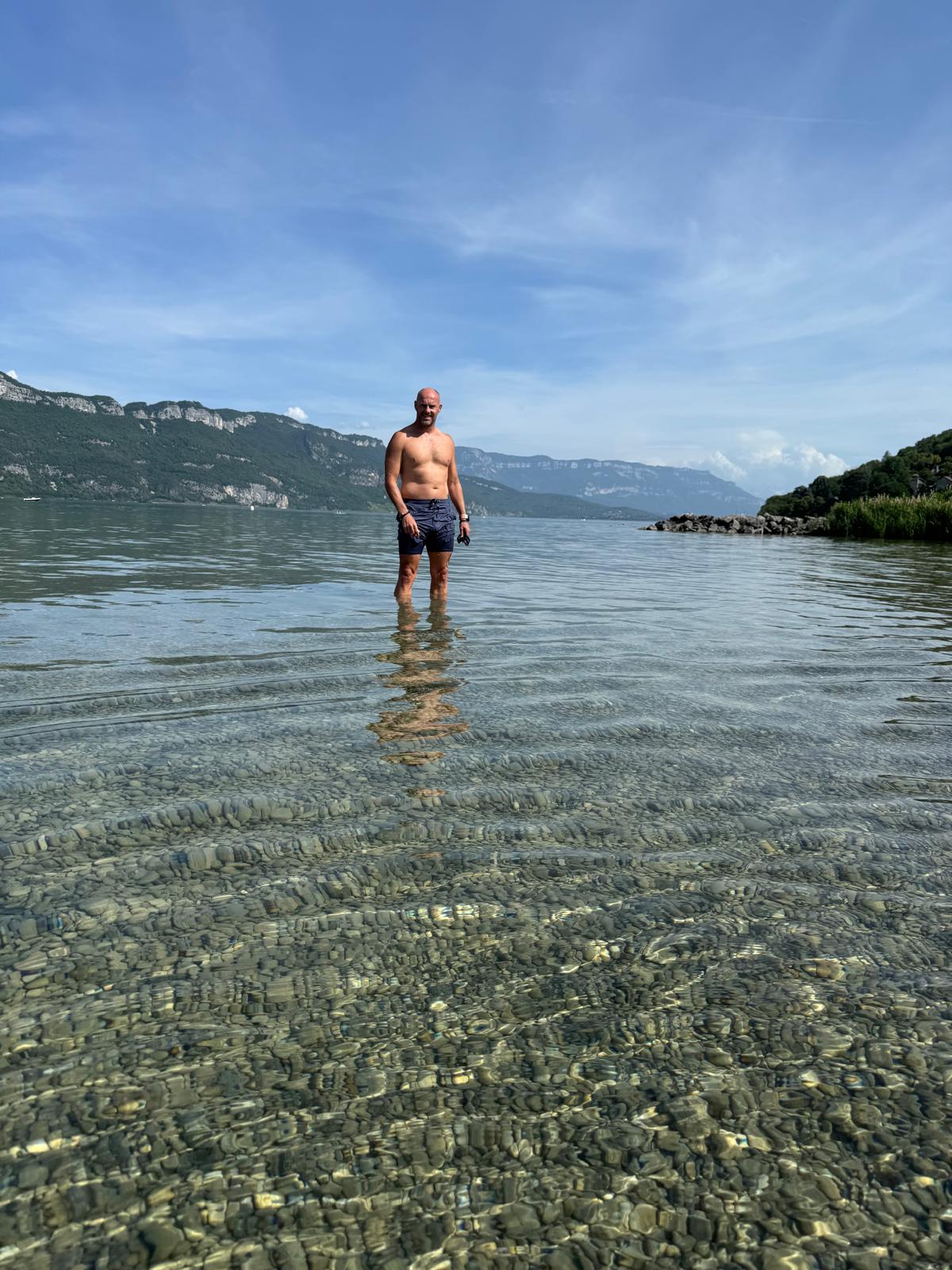 Dave standing waist-deep in the glass-clear water of Lac du Bourget, his reflection on the surface, wooded ridges behind.