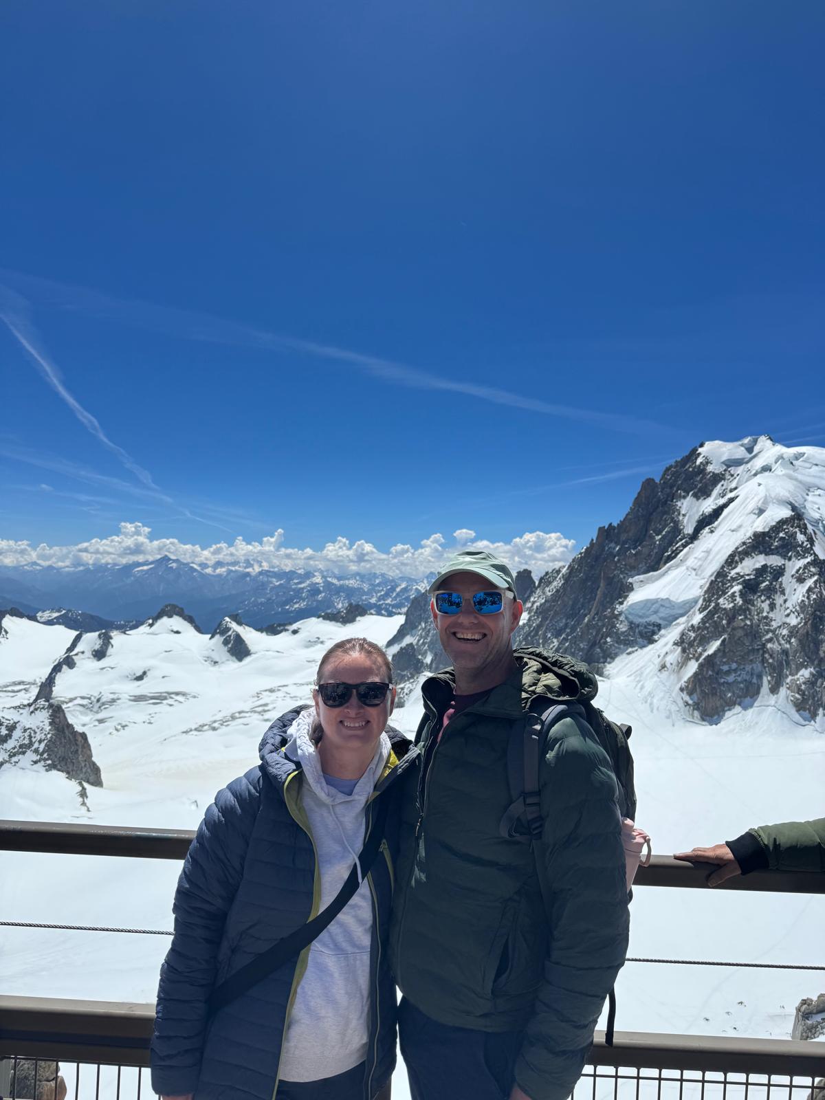 Dave and Lindsey in down jackets at the Aiguille du Midi summit railing, the snow-covered Mont Blanc massif behind.