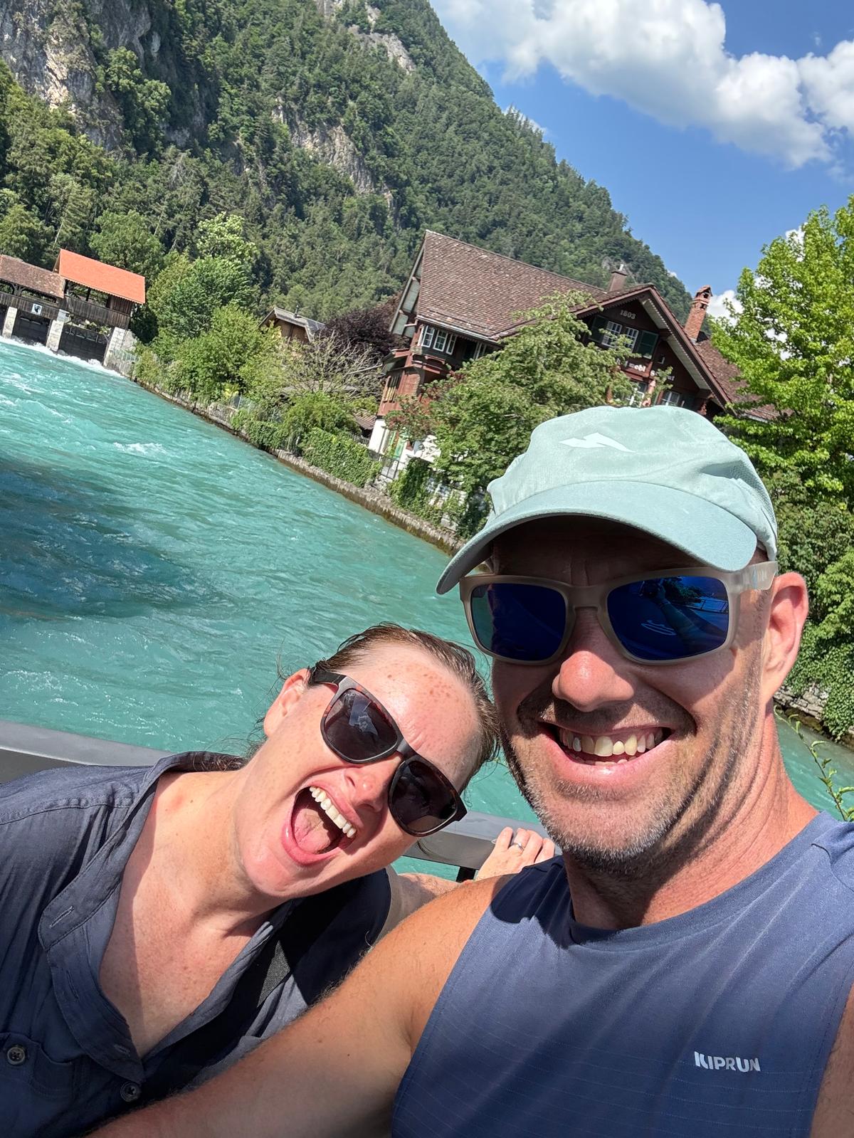 Dave and Lindsey laughing beside the vivid turquoise river Aare at Interlaken, timber chalets and a wooded cliff behind.