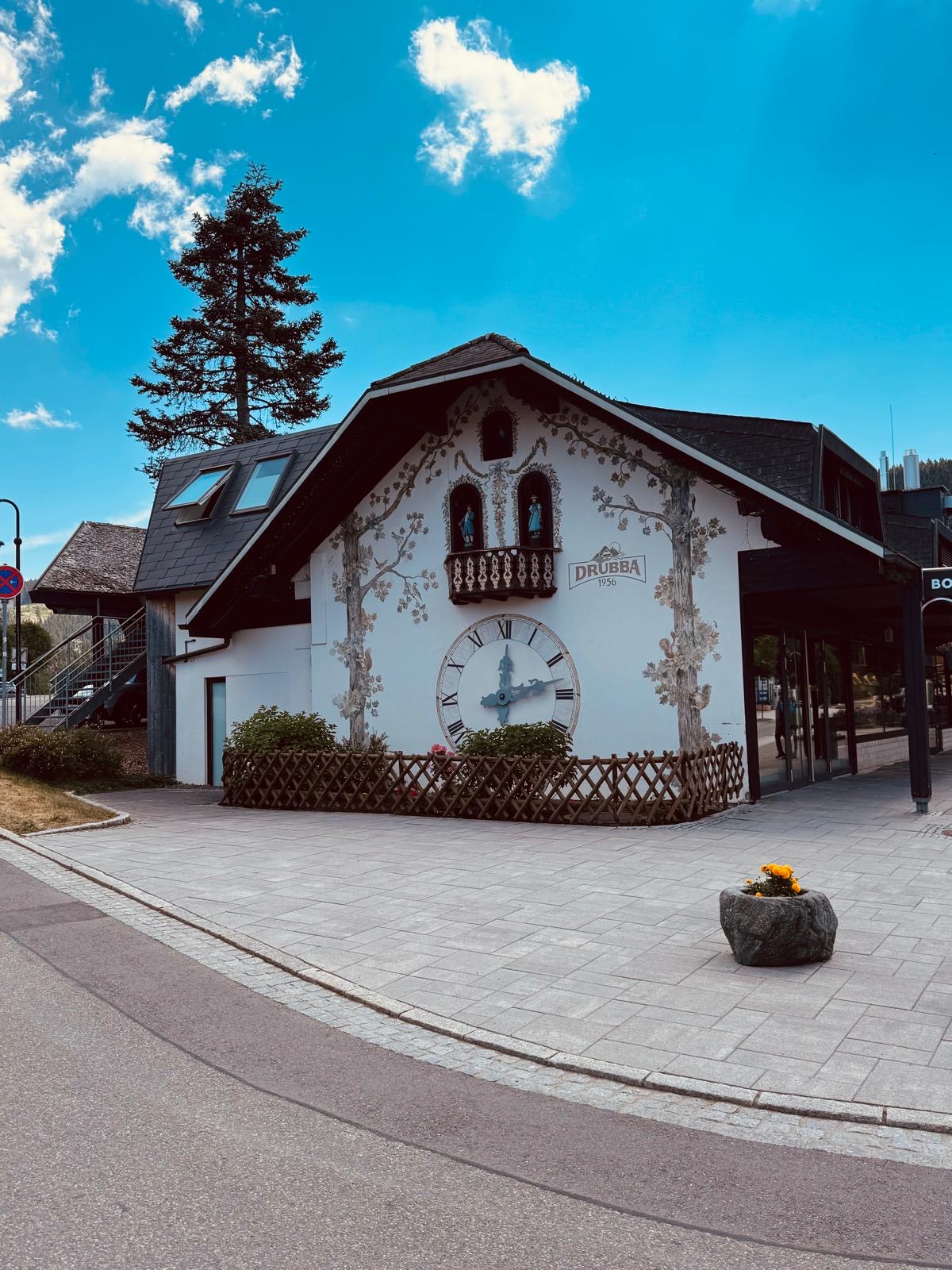 A large white Black Forest building painted to look like a giant cuckoo clock, with a clock face, carved balcony figures and Drubba 1956 signage, at Lake Titisee.