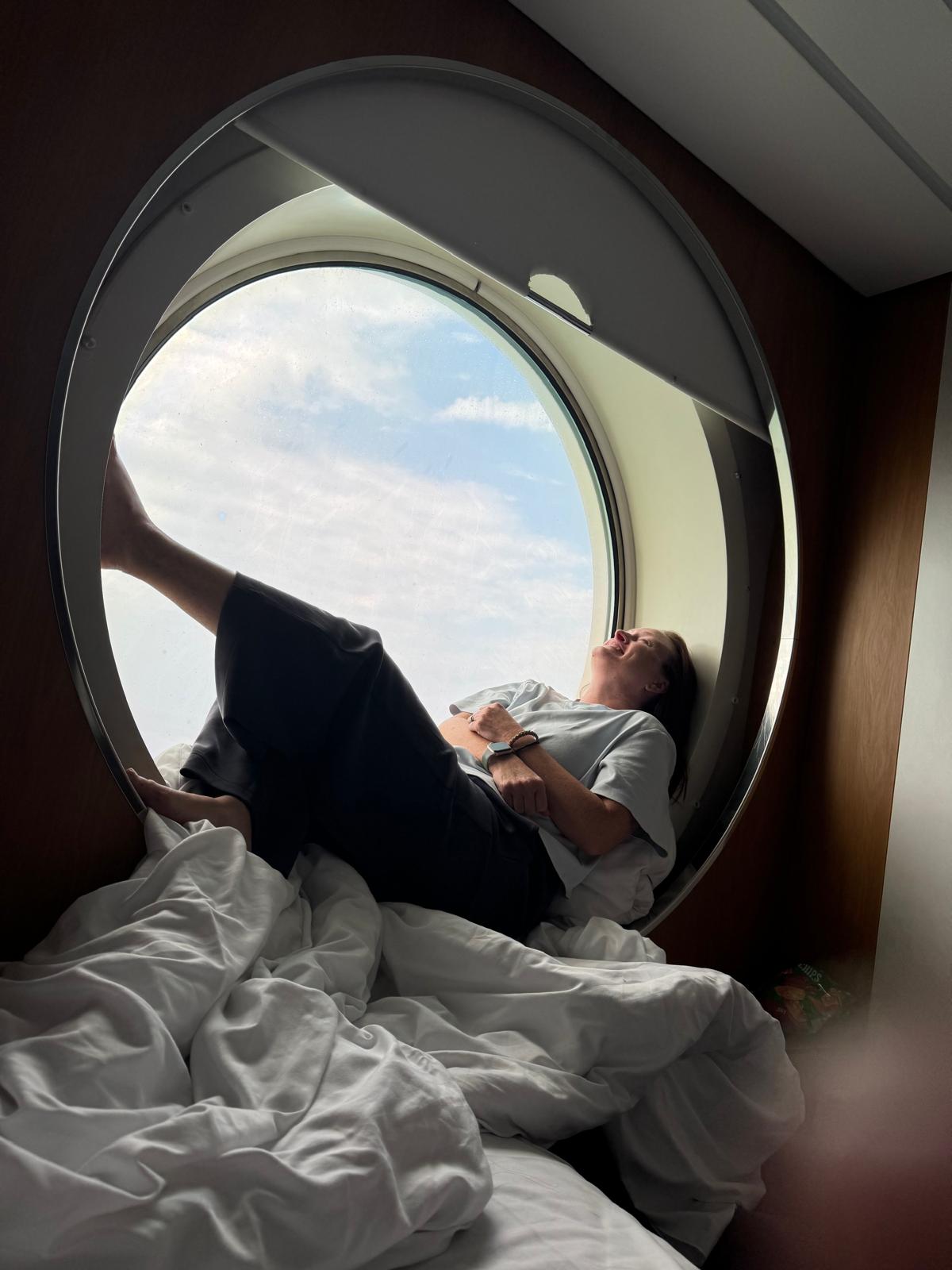 Lindsey lounging in a round ferry-cabin porthole, feet up, laughing, sky through the glass.